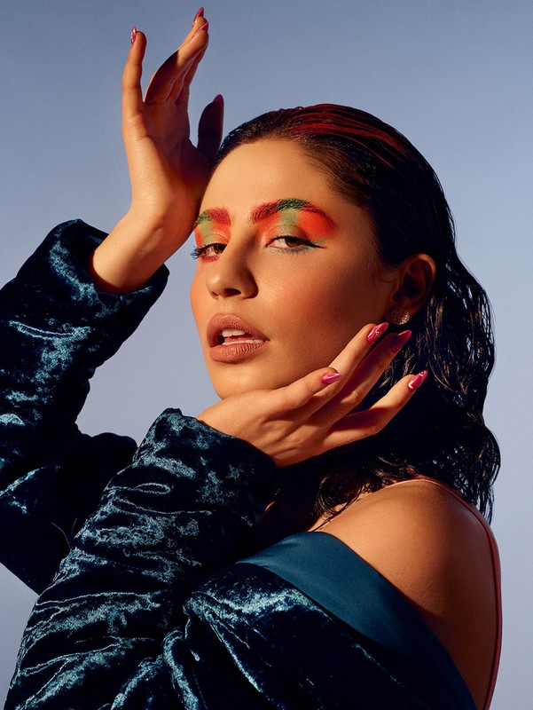 Close-up studio shot of a woman with colorful makeup and a blue velvet jacket, posing with her hands up against a light blue backdrop.