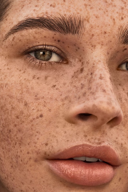 Close-up portrait of a woman with freckled skin, hazel eyes, and glossy lips. The focus is on her face, eyebrows, and complexion.