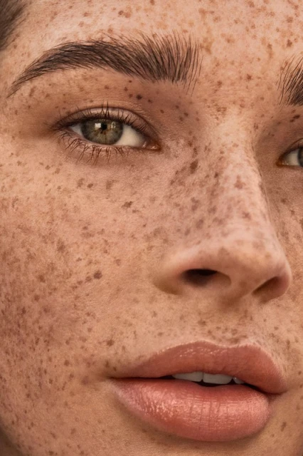 Close-up portrait of a woman with freckled skin, hazel eyes, and glossy lips. The focus is on her face, eyebrows, and complexion.