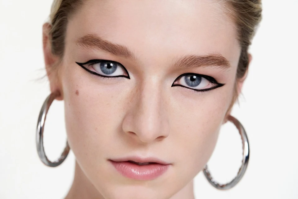 Close-up portrait of an adult woman with blue eyes and graphic black eyeliner, complemented by large silver hoop earrings.