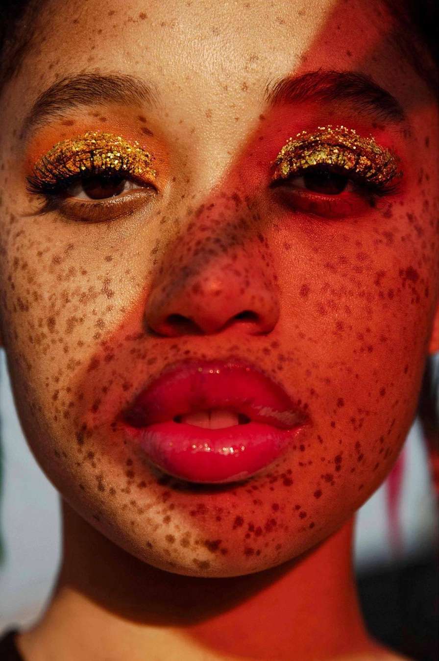 Close-up portrait of a freckled young woman with gold glitter eye makeup and glossy red lips under split red and warm lighting.
