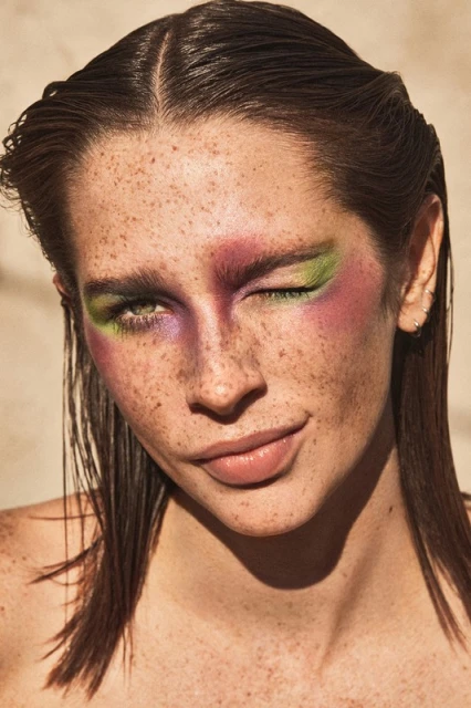 Close-up shot of a young adult woman with freckles, wearing vibrant makeup with a winking expression, and wet hair in an indoor setting.