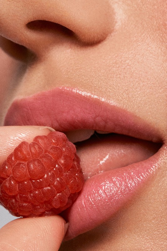 Macro shot of a woman's face showing lips, teeth, finger, and raspberry, creating a beauty or food-related visual.