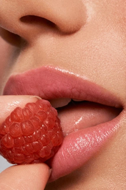 Macro shot of a woman's face showing lips, teeth, finger, and raspberry, creating a beauty or food-related visual.
