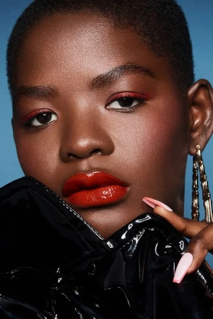 Close-up studio portrait of a Black woman with glossy red lips, matching red eyeliner, gold hoop earrings, and a shiny black jacket.
