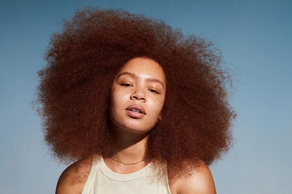 Close-up portrait of a young adult woman with natural, voluminous auburn afro hair, tan skin, and a neutral facial expression against a blue backdrop.