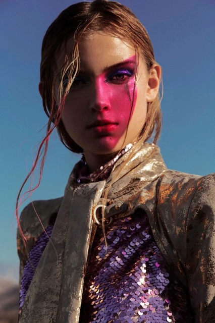 Close-up portrait of a young woman with creative pink and purple makeup, wearing a sequined top and gold jacket against a clear blue sky.