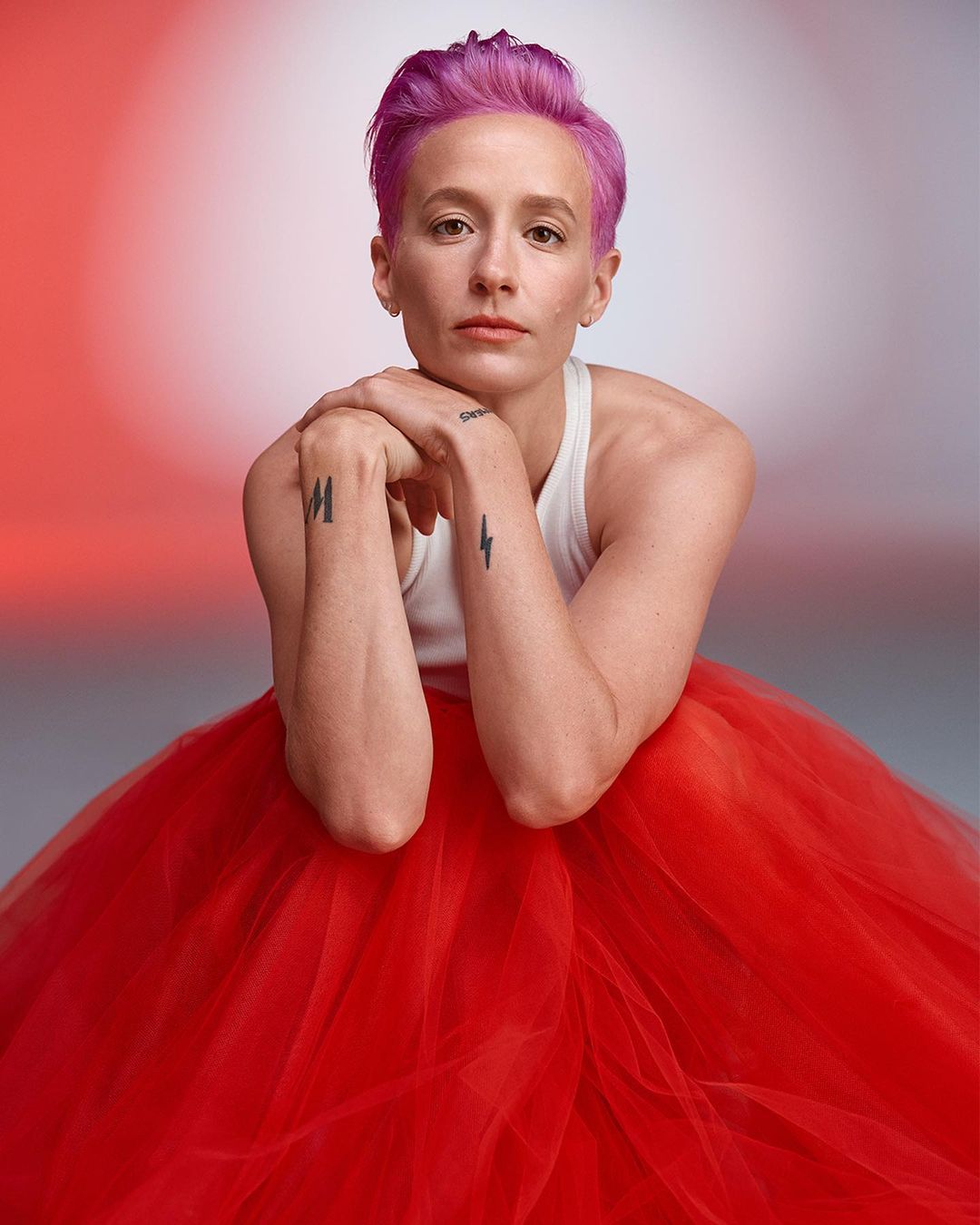 Portrait of an adult female with short pink hair, wearing a white top and a large red tulle skirt, with a neutral facial expression.