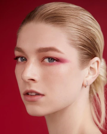 Close-up portrait of a woman with slicked-back blonde hair and dramatic pink eyeshadow against a red background.
