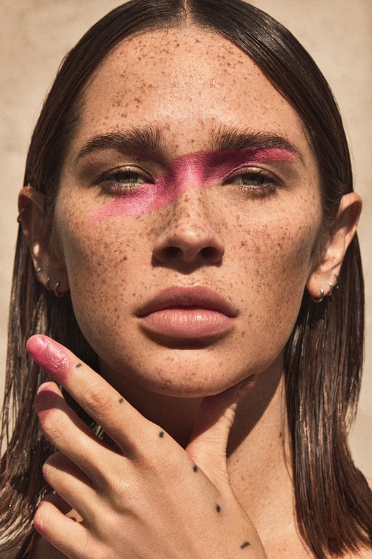 Portrait of a young woman with pink face paint and freckles, hand near her neck, medium-shot in front of a neutral background.