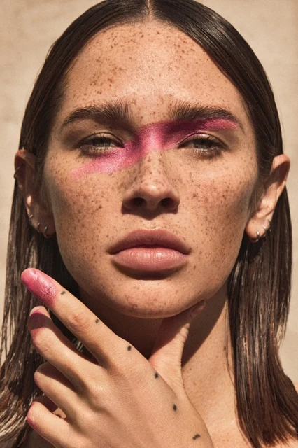 Portrait of a young woman with pink face paint and freckles, hand near her neck, medium-shot in front of a neutral background.