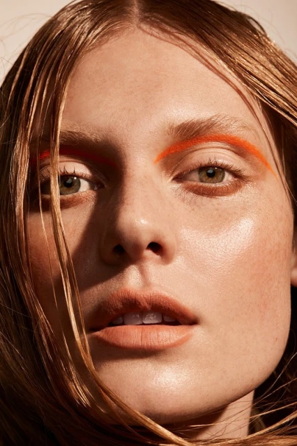 Close-up portrait of a woman with wet blonde hair and striking orange makeup on her eyebrows in a beauty or fashion context.