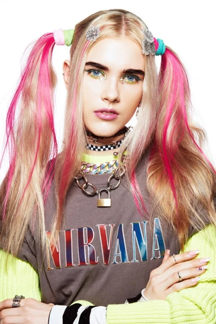 Blonde woman with pigtails, vibrant makeup, and vintage fashion stands in a photo studio, looking at the camera.