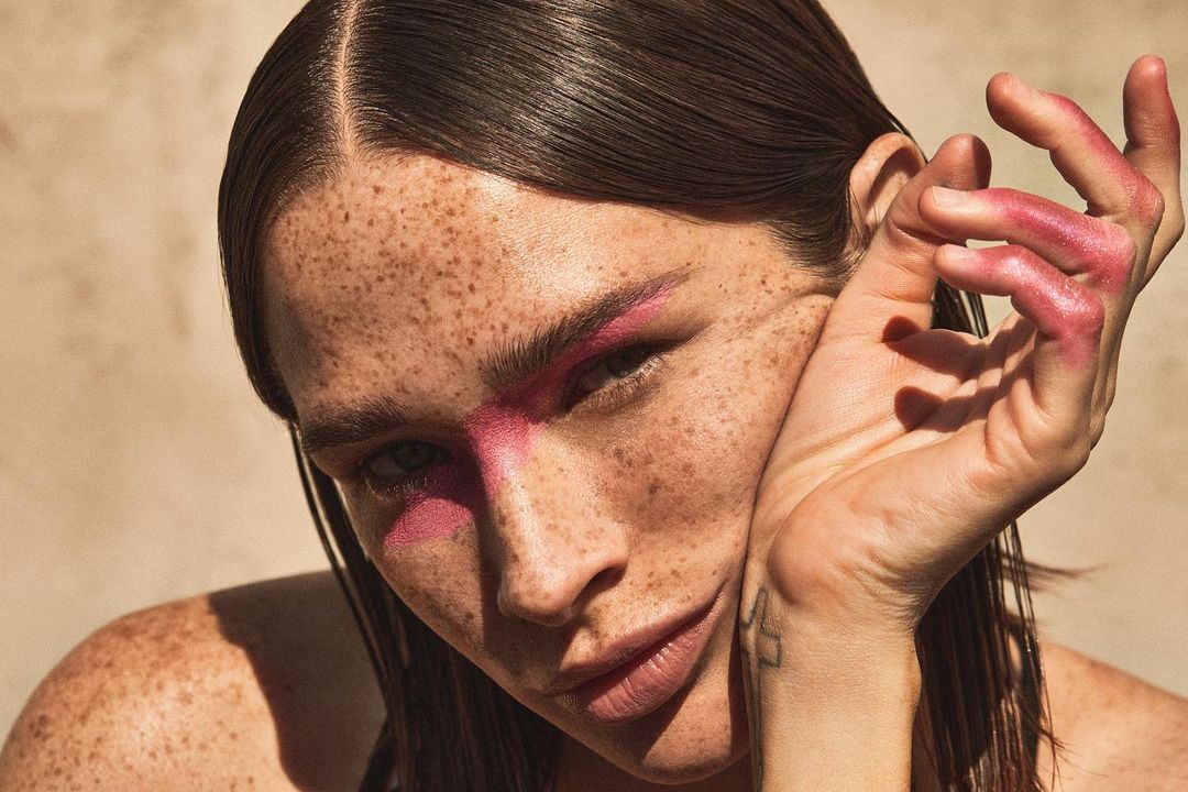 Close-up of an adult woman with freckles and pink makeup on her face, posing with her hand near her face in a beauty shot.