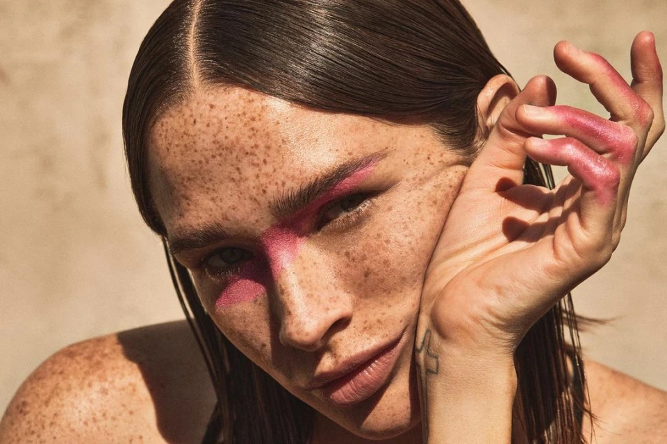 Close-up of an adult woman with freckles and pink makeup on her face, posing with her hand near her face in a beauty shot.