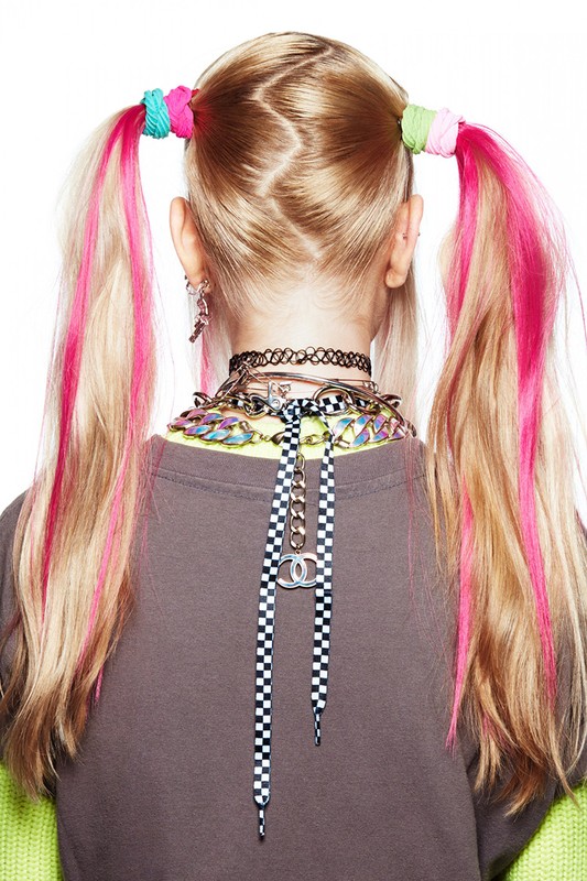 Back view of a child with blonde hair in pigtails, accessorized with colorful scrunchies and multiple necklaces against a white background.