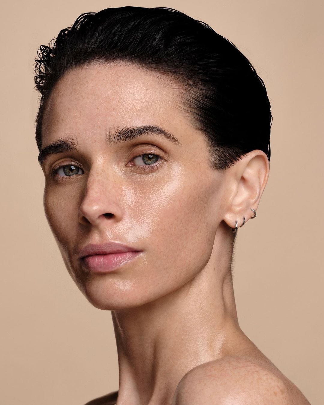 Close-up studio shot of a fair-skinned adult woman with short dark hair and hoop earrings, gazing intently at the camera against a tan backdrop.