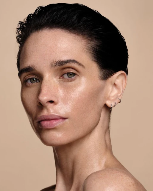 Close-up studio shot of a fair-skinned adult woman with short dark hair and hoop earrings, gazing intently at the camera against a tan backdrop.
