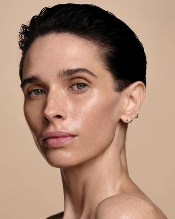 Close-up studio shot of a fair-skinned adult woman with short dark hair and hoop earrings, gazing intently at the camera against a tan backdrop.