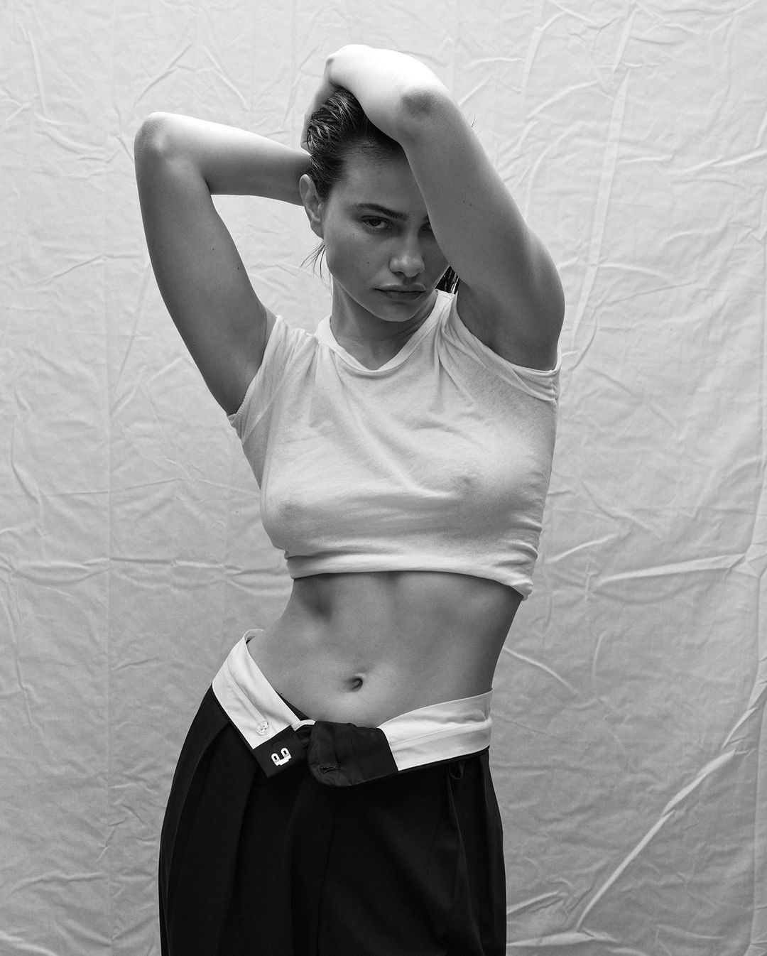 Black and white studio portrait of a young adult woman in a crop top and pants, posing with her hands behind her head, looking at the camera.