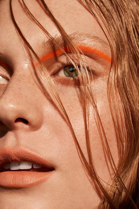 Close-up studio portrait of a young woman with red hair and orange eyeliner, showcasing beauty and fashion trends with soft lighting.