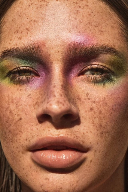 Close-up portrait of a woman with freckles and colorful eye makeup including pink, green, and purple eyeshadow. High-key lighting.