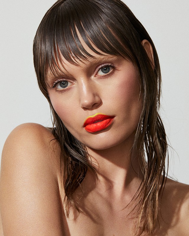 Close-up portrait of a woman with wet dark hair, light skin, light eyes, and orange lipstick with yellow outlining against a light background.