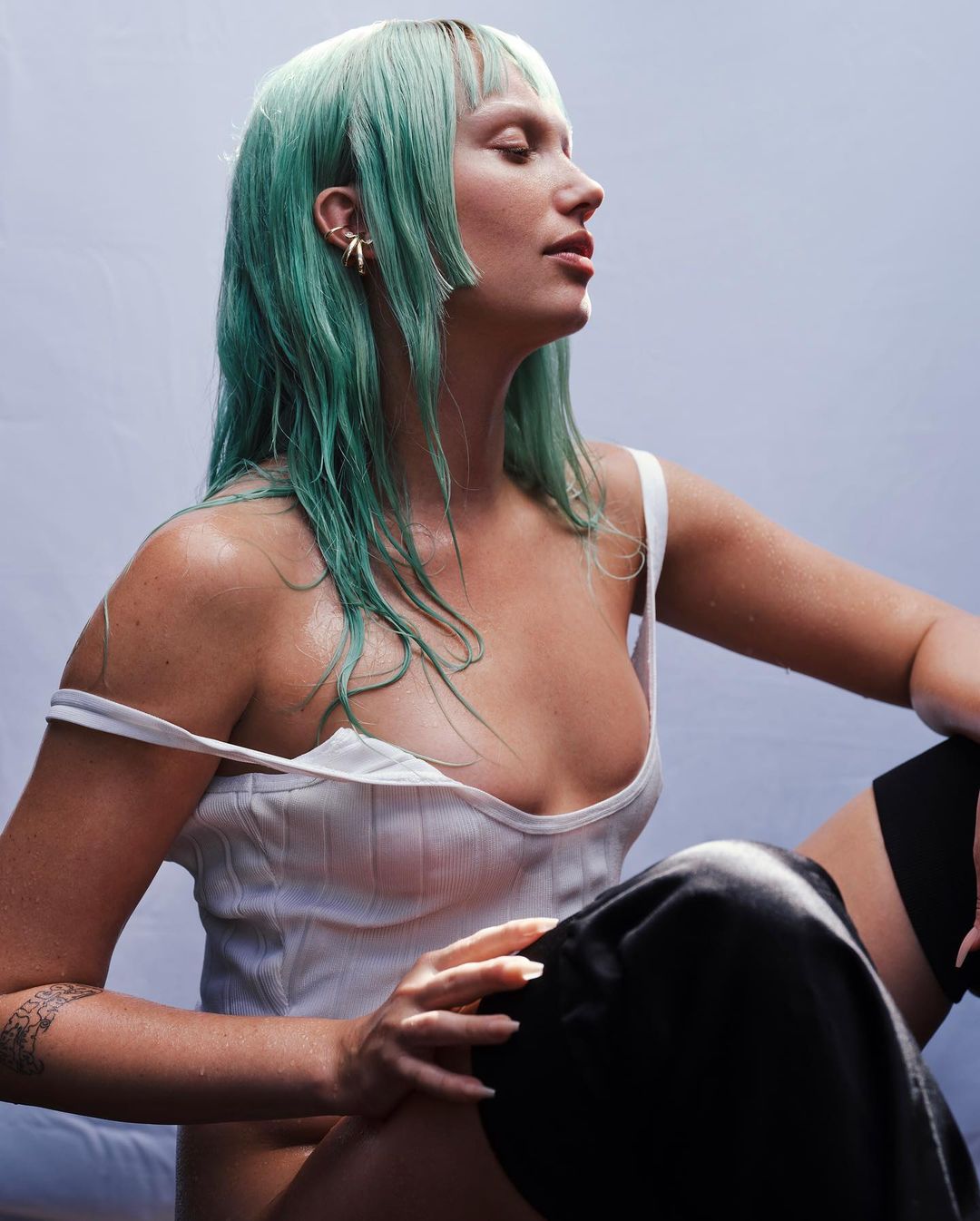 Portrait of woman with wet, green hair wearing a white tank top. She's sitting with one leg up, shot in a studio setting against a light backdrop.