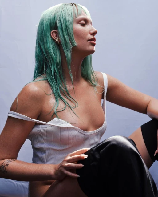 Portrait of woman with wet, green hair wearing a white tank top. She's sitting with one leg up, shot in a studio setting against a light backdrop.