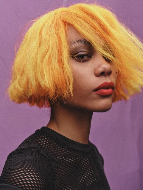 Portrait of a young adult female with bright yellow, short hair, red lipstick, wearing a black mesh shirt, in a photo studio with purple background.