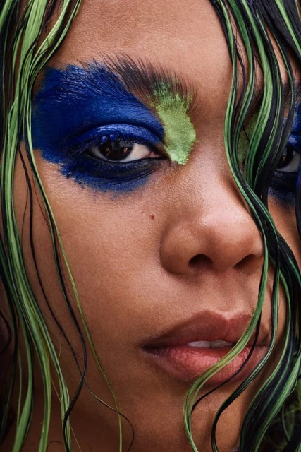 Close-up portrait of a woman with dramatic blue and green eye makeup and wet black and green hair framing her face.