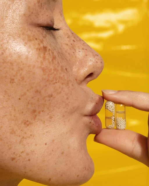 Close-up of a fair-skinned adult female with freckles. Two clear capsules filled with white beads are held between her lips and fingers.