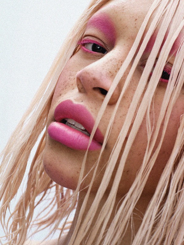 Close-up of a young adult woman with bright pink eyeshadow and lipstick, light blonde hair partially obscures her face.