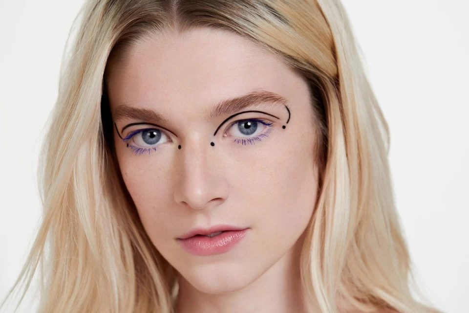 Close-up portrait of a fair-skinned woman with blonde hair and blue eyes, wearing decorative black and violet eyeliner, on a white backdrop.