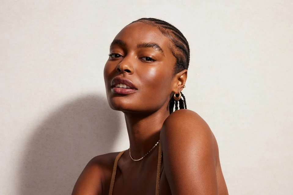Portrait of an adult woman with dark skin and braided hair, wearing a gold necklace, looking up against a light beige backdrop.