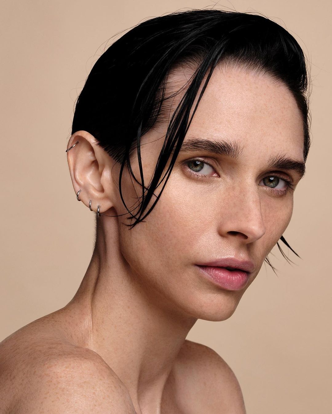 Close-up beauty portrait of a young adult woman with dark wet hair, bare shoulders, a neutral expression, and minimal makeup on a neutral background.