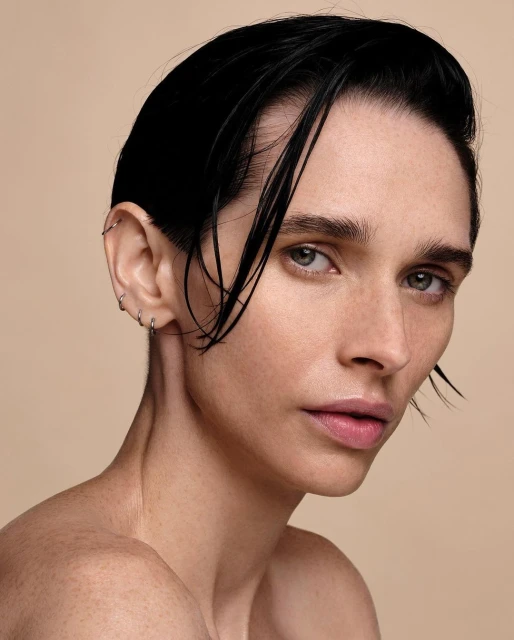 Close-up beauty portrait of a young adult woman with dark wet hair, bare shoulders, a neutral expression, and minimal makeup on a neutral background.