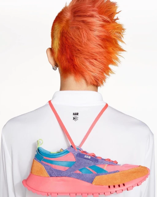 Person with vibrant orange hair displaying a colorful Reebok sneaker from behind, worn as a neck accessory against a white backdrop.