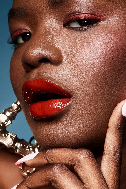 Close-up shot of a black female model with red eye makeup and glossy red lips wearing large gold hoop earrings on a blue background.