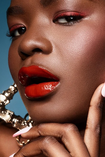 Close-up shot of a black female model with red eye makeup and glossy red lips wearing large gold hoop earrings on a blue background.