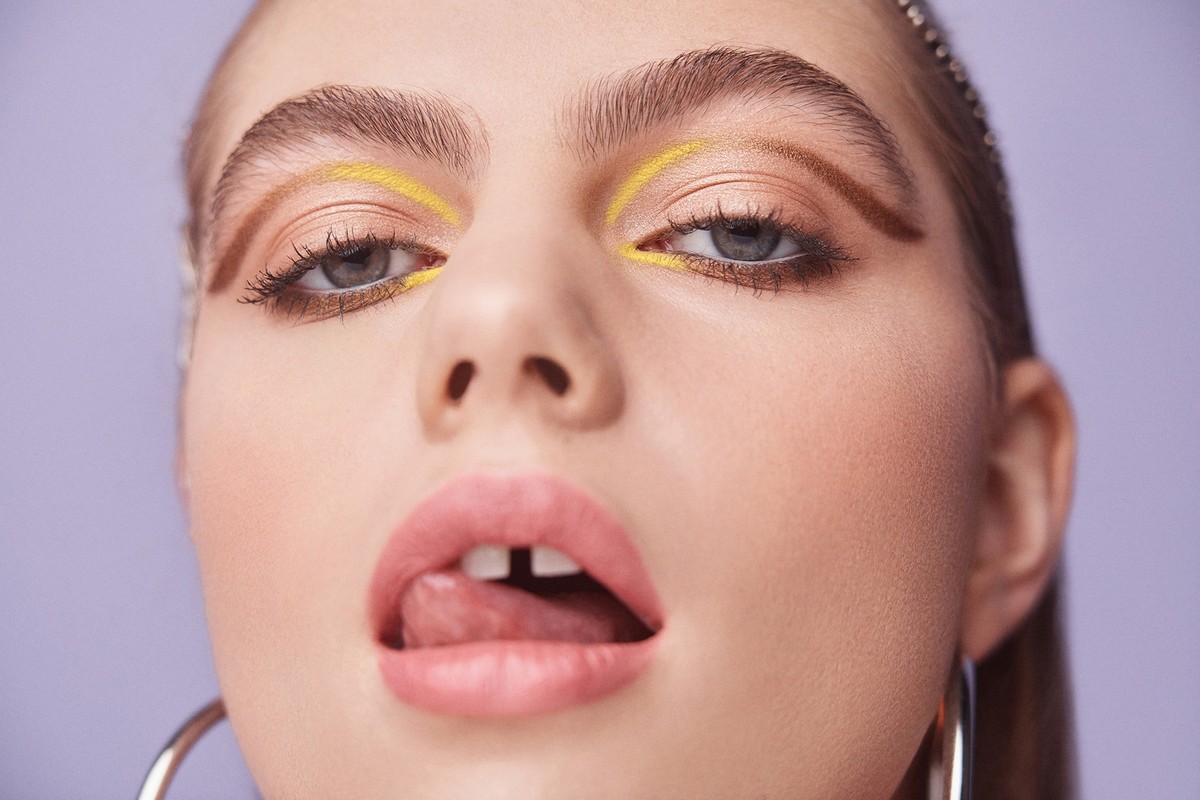 Close-up portrait of a young woman with unique makeup, including yellow eyeliner and bold brows, set against a solid light purple backdrop.