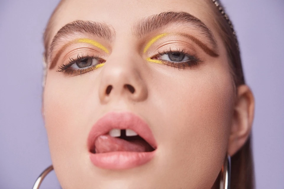 Close-up portrait of a young woman with unique makeup, including yellow eyeliner and bold brows, set against a solid light purple backdrop.