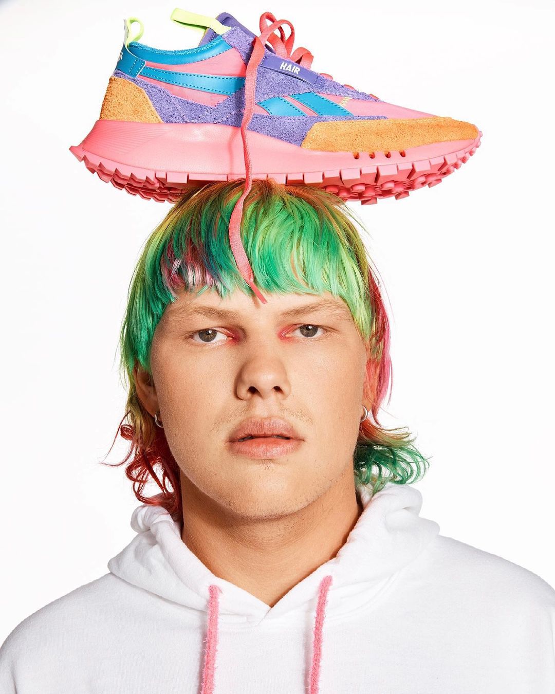 Person with colorful hair wearing a white hoodie and balancing a pink, blue, and orange Reebok sneaker on their head.
