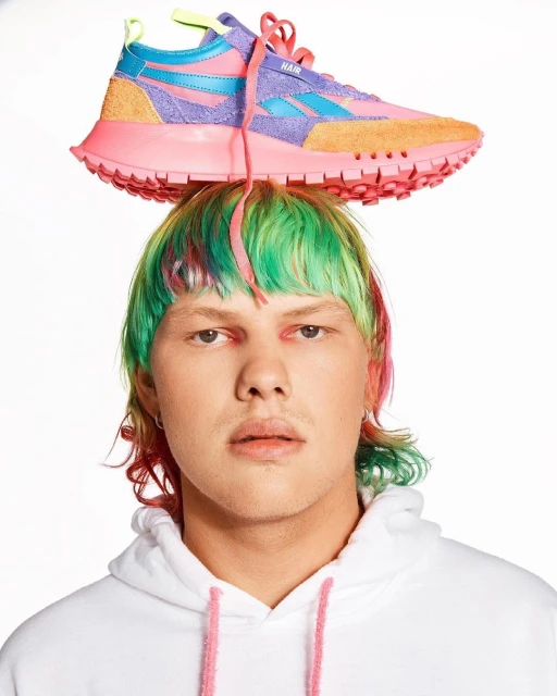 Person with colorful hair wearing a white hoodie and balancing a pink, blue, and orange Reebok sneaker on their head.