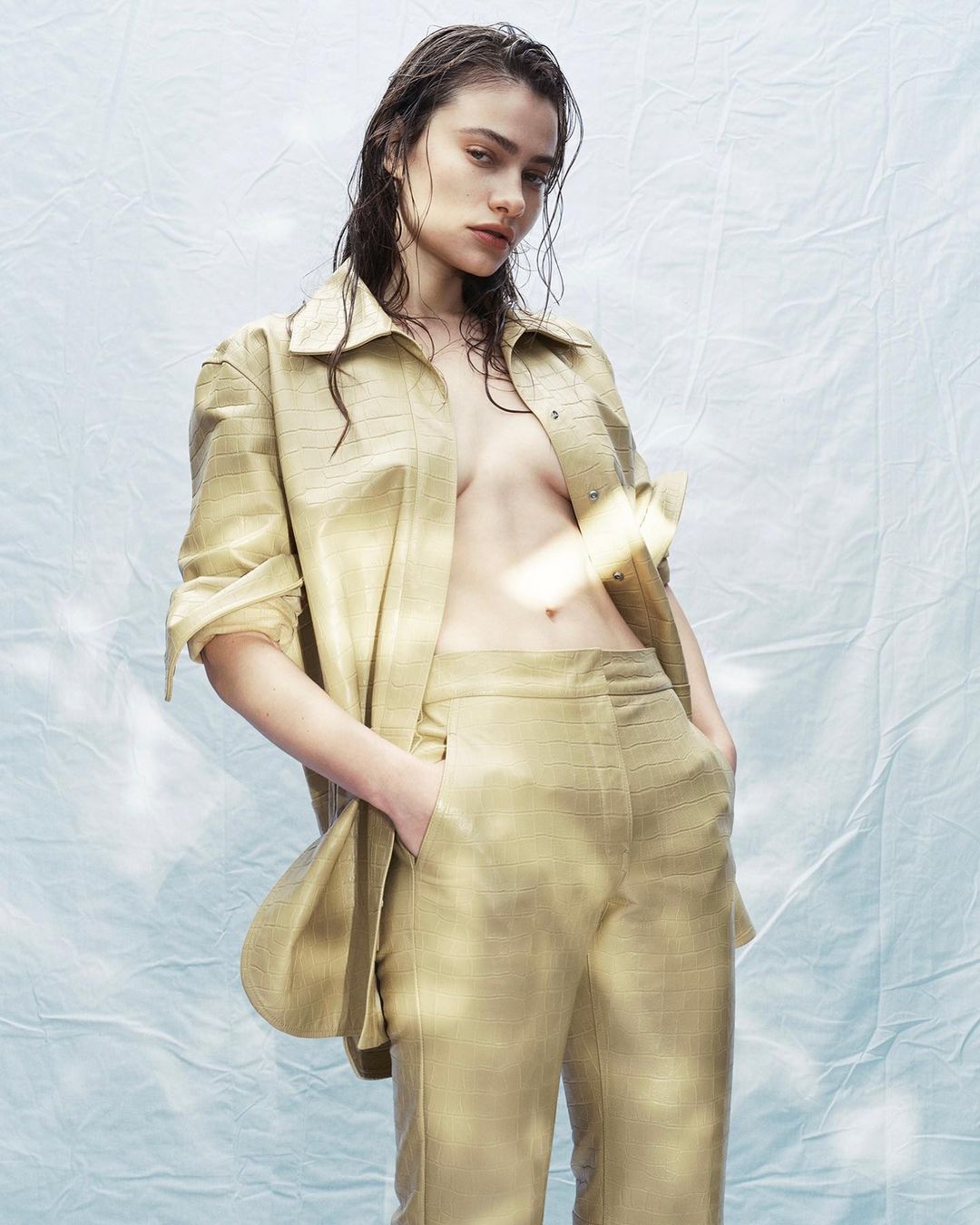 Woman in a tan-colored, collared shirt and matching pants poses indoors with wet hair against a mottled light blue and white backdrop.