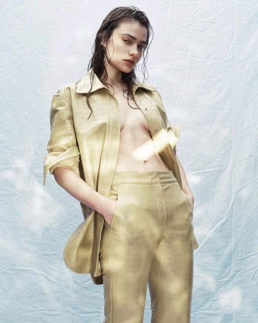Woman in a tan-colored, collared shirt and matching pants poses indoors with wet hair against a mottled light blue and white backdrop.