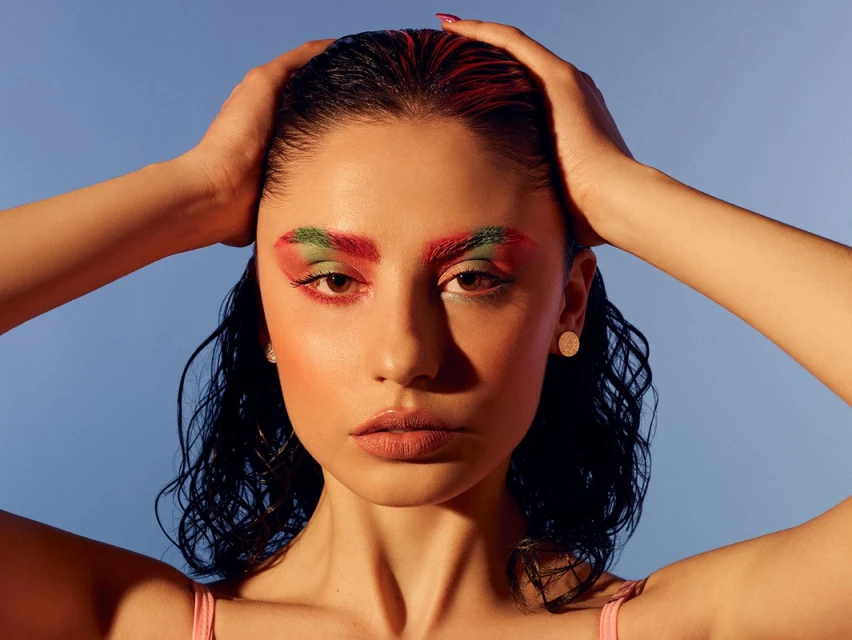 Close-up portrait of a woman with wet hair, striking colorful eye makeup, and hands on her head, set against a clear blue background.