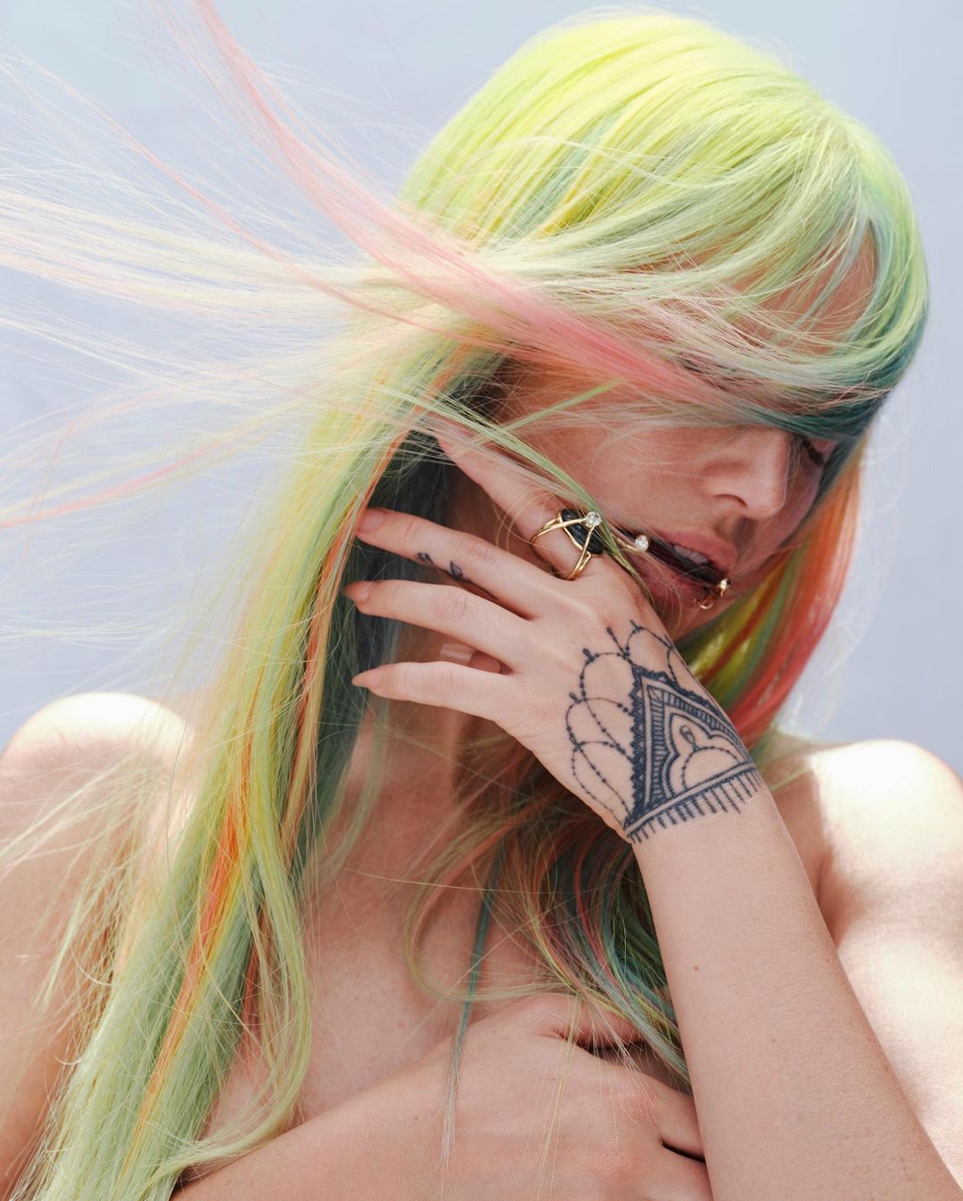 Portrait of a person with rainbow-colored hair, hand tattoo, and jewelry, posing indoors, against a bright backdrop.