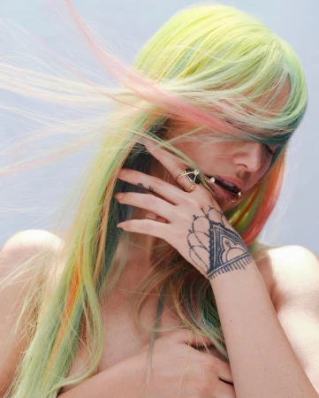 Portrait of a person with rainbow-colored hair, hand tattoo, and jewelry, posing indoors, against a bright backdrop.