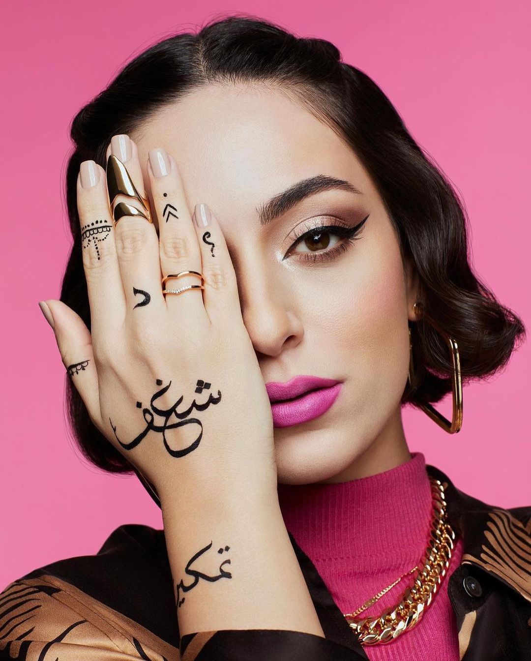 Close-up shot of a woman with henna tattoos on her hand, statement rings, pink lipstick, and a pink background for a beauty ad.